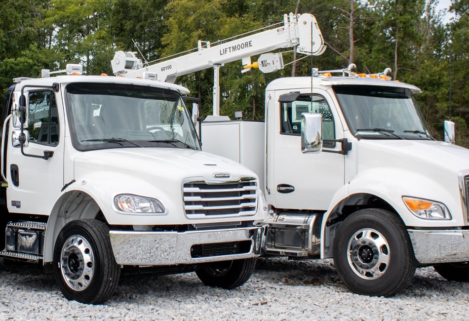 Two white trucks with aerial lifts on a gravel surface with trees in the background